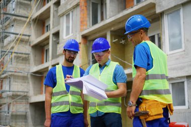 Three construction workers reviewing blueprints outside unfinished building