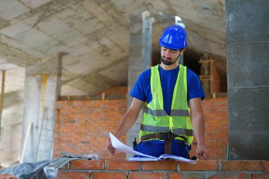 Güvenlik yeleği giyen erkek inşaat işçisi bina planlarını inceliyor.