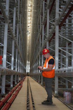 Depo raflarında turuncu güvenlik yeleği giyen bir adam var.