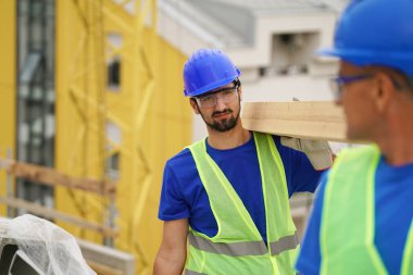 Construction worker carrying wooden plank wearing safety gear outdoors