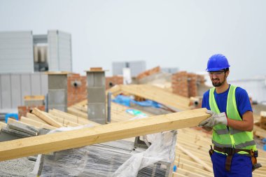 İnşaat işçisi inşaat alanında ahşap kiriş taşıyor.