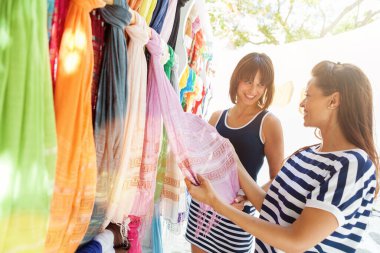 İki kadın dışarıda alışveriş yapıyor, renkli asılı eşarpları inceliyor, mutlu bir şekilde gülümsüyor.