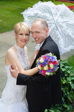 Gülümseyen gelin ve damat ellerinde buket şemsiyeyle dışarıda poz veriyorlar.
