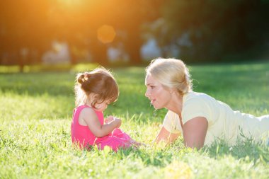Gülümseyen kadın yeşil çimlerde oynayan genç kızın yanında yatıyor.