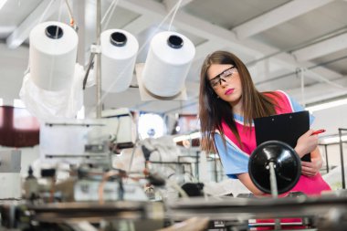 Kapalı alanda pano tutarken tekstil makinelerini inceleyen kadın