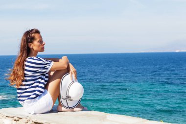 Mavi okyanusun kenarında oturan genç bir kadın elinde beyaz şapkayla dinleniyor.