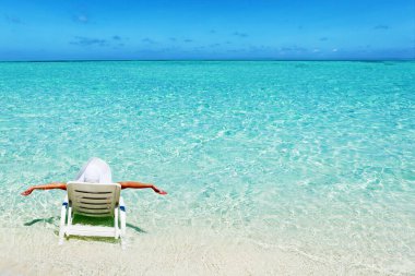 Genç bir kadın tropik plajda güverte sandalyesinde dinleniyor. 