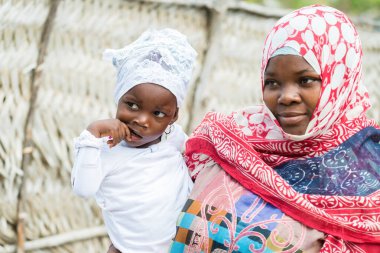 Gülümseyen bir kadın açık havada renkli giysiler içinde küçük bir çocuğu tutuyor.