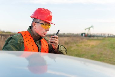 Turuncu yelek giyen erkek işçi açık havada telsiz kullanıyor.
