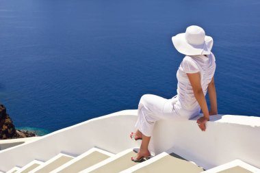 Yunanistan 'ın Santorini kentinde mavi okyanus manzarasının yanında dinlenen beyaz şapkalı kadın
