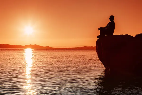 Bir adamın silueti denizin üstündeki taşın üzerinde meditasyon yapar, gün batımı ışığı.