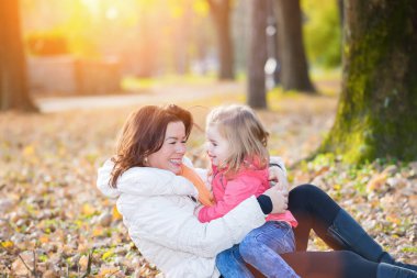 Gülümseyen kadın, güz yapraklarının arasında oturan genç kızı kucaklıyor.