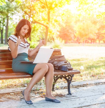 Parkta dizüstü bilgisayar kullanan mutlu genç bir kadın.