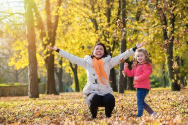 Anne ve küçük kızı sonbahar parkında yapraklarla oynuyorlar.