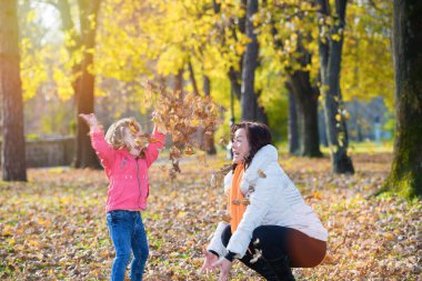 Anne ve küçük kızı sonbahar parkında yapraklarla oynuyorlar.