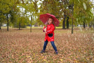 Yağmurluklu ve şemsiyeli küçük kız parkta oynuyor.  