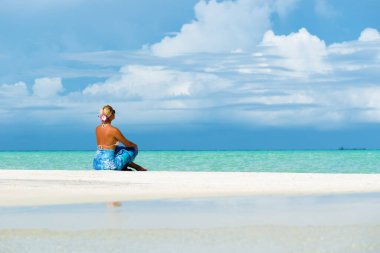 Sarışın kadın beyaz kumsalda oturmuş açık denize bakıyor.