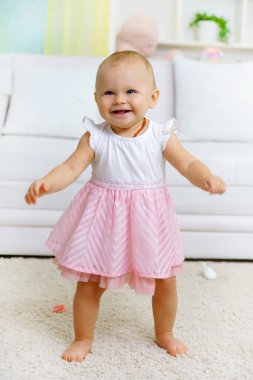 Küçük tatlı kız kanepenin yanında yürüyor, kameraya bakıyor.                                   