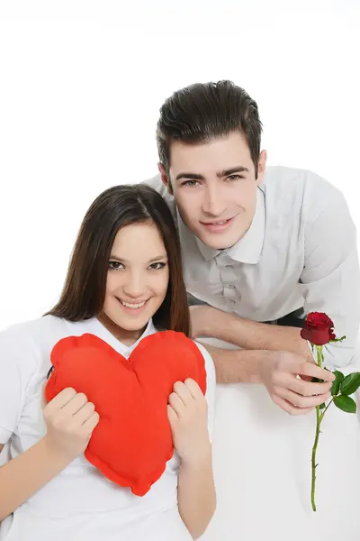 Güzel, kalp şeklinde yastığı olan genç bir kadın, arkasında duran ve elinde kırmızı bir gül çiçeği tutan bir adam.