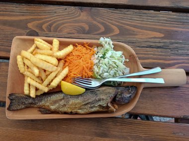 Tahta tabakta servis edilen kızarmış alabalık, patates kızartması ve salata.