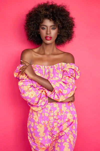 自信を持ってアフリカ系アメリカ人女性で流行の服とアフリカの髪型でカメラを見ながら スタジオでピンクの背景を背景に立って — ストック写真