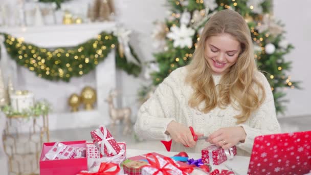 Młoda Blondynka Pakująca Pudełko Świąteczne Siedząca Obok Stołu Salonie Sosną — Wideo stockowe