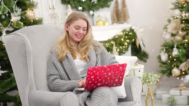 一个微笑的年轻女人的画像 她抱着笔记本电脑在她的小腿上工作 坐在圣诞树旁舒适的扶手椅上 — 图库视频影像