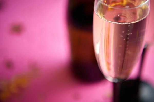 Bottle and two glasses full of sparkling champagne wine with golden decoration of confetti and serpentines on pink background. Top view with copy space. Festive or party concept.