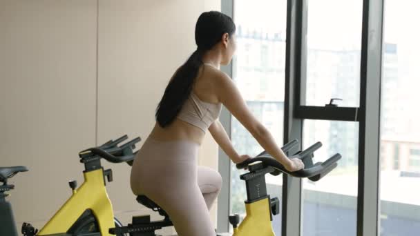 Молодая Азиатка Тренируется Стационарном Велосипеде Крытом Тренажерном Зале Окруженном Окнами — стоковое видео