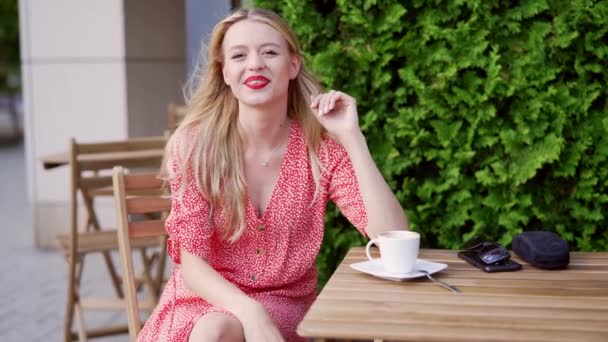 穿着时髦衣服的金发美女坐在餐桌边 在街上的咖啡馆里 一边摸着脖子一边笑 — 图库视频影像