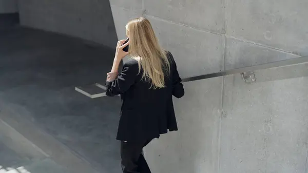 一个穿着黑色西服的金发女商人 穿着现代混凝土建筑 一边打电话一边聊天 — 图库照片