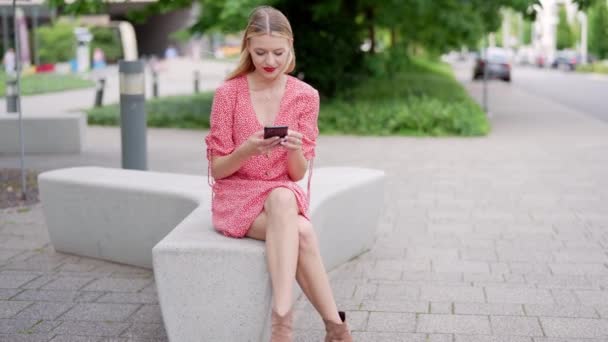 ファッショナブルな赤いドレスの若い女性は 静かな都市公園の緑に囲まれた彼女の携帯電話を使用してベンチに座っています — ストック動画