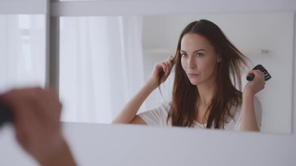 一位年轻女子一边在明亮的房间里看着镜子 一边用卷发钳梳理头发 这反映出她的美感和仪态整洁 — 图库视频影像