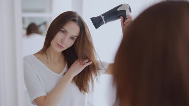 カジュアルなTシャツの女性は明るく照らされた部屋の鏡の前で吹くドライヤーを使用して髪をスタイルします — ストック動画