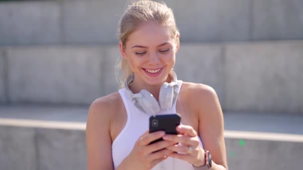 在阳光明媚的日子里 一个戴着耳机的年轻貌美的女人在户外用她的智能手机 — 图库视频影像