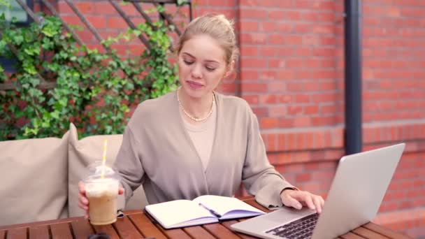 专注的女商人在一家舒适的户外咖啡馆里用笔记本电脑 用吸管喝水 — 图库视频影像