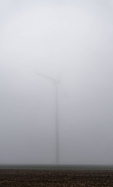 Kış günü siste gizlenmiş tarlada zar zor görülebilen rüzgar türbini.