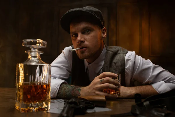 Cards and revolver lying on table in front of dissatisfied young man in waistcoat and flat cap smoking, drinking whiskey and  looking defiantly at camera. Vintage look inspired by Peaky Blinders