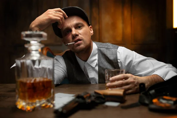 Young tipsy man in white shirt, vest and peaked flat cap sitting with glass of whiskey at table in vintage casino with cards and pistol lying nearby, smoking cigarette and looking defiantly at camera