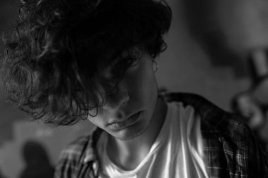 Young guy looking defiantly at camera through dark curly hair falling over his eyes. Closeup portrait in black and white. Modern youth lifestyle concept