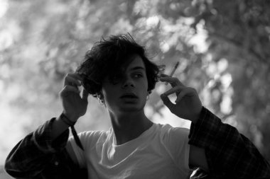 Relaxed young guy with tousled dark hair in white t-shirt and plaid shirt holding half-smoked cigarette and looking away indifferently  while standing in shadow of trees. Black and white shooting
