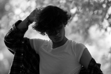 Relaxed guy with head down and dark wavy hair falling over face in white t-shirt and plaid shirt pulled down from shoulders standing outdoors on blurred background of tree leaves. Black and white shot