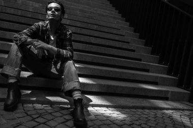 Relaxed confident young guy posing on stone stairs on city street, looking defiantly at camera through sunglasses. Zoomers generation concept. Urban lifestyle