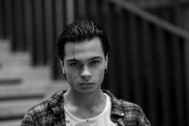 Handsome slightly unshaven guy with styled dark hair and piercing in ears calmly and confidently looking at camera while standing on blurred background of urban construction. Black and white portrait