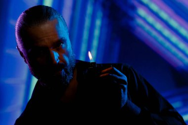 Confident handsome bearded man standing in dark of night city on background of blue neon lighting, holding lit cigarette lighter