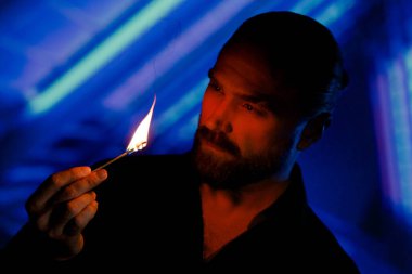 Portrait of confident relaxed handsome bearded man standing in dark of night city on background of blue neon lighting, looking at lit match in his hand