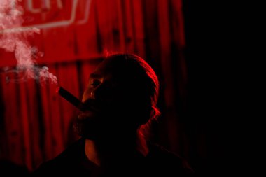 Relaxed bearded man enjoying fragrant cigar in darkness, leisurely smoking with closed eyes to take mind off everyday concerns