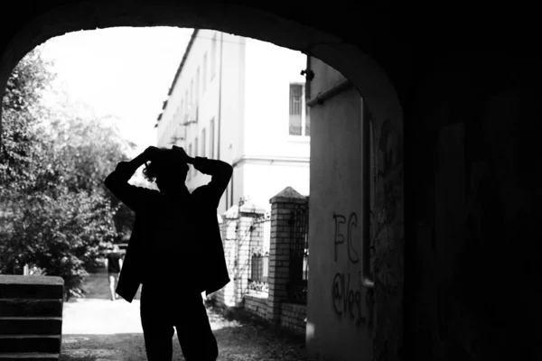 Young guy with face hidden in shadows standing in house archway with graffiti painted walls on background of sunny summer city street. Black and white silhouette photography