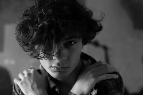 Closeup black and white portrait of modern urban teenager with piercing in ear and dark wavy hair falling over face posing with hands on his shoulders, looking defiantly at camera