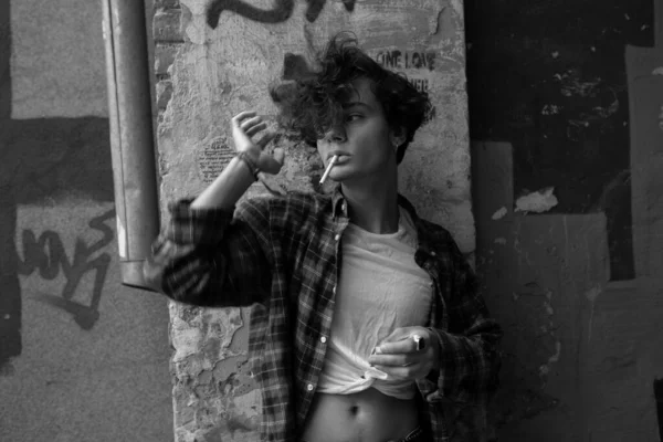 Relaxed young guy standing with cigarette in mouth leaning against graffiti painted cement wall of old building, adjusting his black curly hair and looking away. Black and white fashion photo